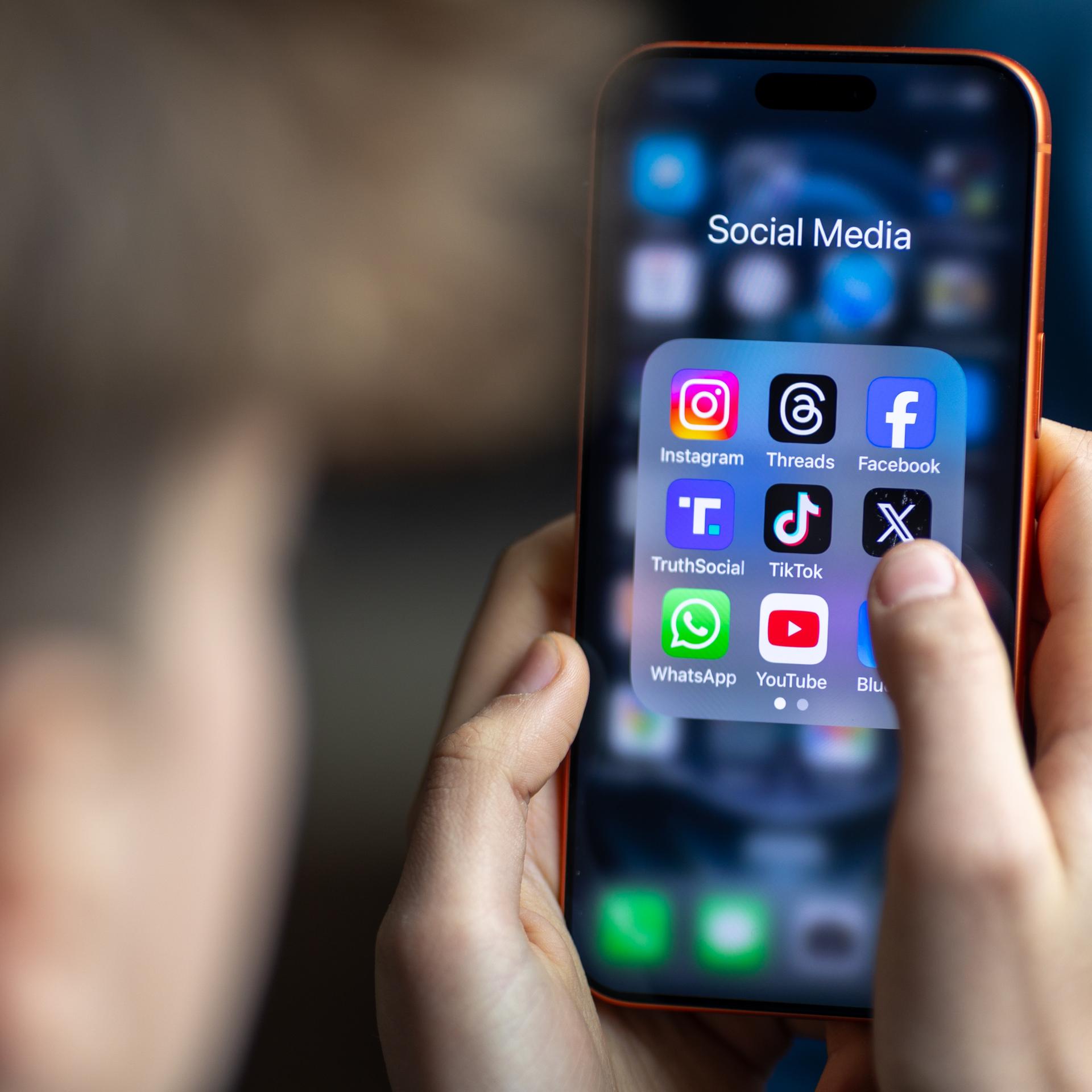 Ein Jugendlicher mit einem Smartphone auf dessen Display u.a. viele Social Media Anwendungen von Meta zu sehen sind. Der Hintergrund ist dunkel und verschwommen. Vom Jugendlichen ist nur ein Profil und die Hände zu sehen. Der Fokus ist auf dem Smarphone.