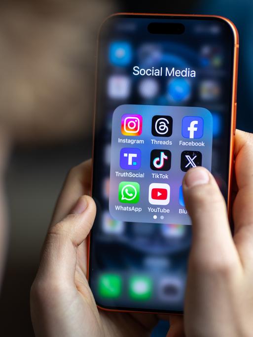 Ein Jugendlicher mit einem Smartphone auf dessen Display u.a. viele Social Media Anwendungen von Meta zu sehen sind. Der Hintergrund ist dunkel und verschwommen. Vom Jugendlichen ist nur ein Profil und die Hände zu sehen. Der Fokus ist auf dem Smarphone.