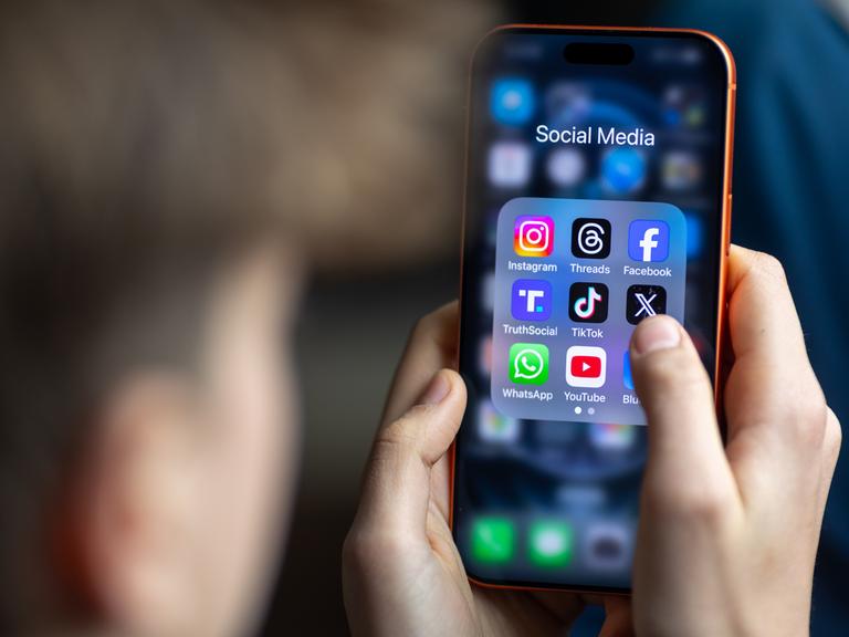 Ein Jugendlicher mit einem Smartphone auf dessen Display u.a. viele Social Media Anwendungen von Meta zu sehen sind. Der Hintergrund ist dunkel und verschwommen. Vom Jugendlichen ist nur ein Profil und die Hände zu sehen. Der Fokus ist auf dem Smarphone.