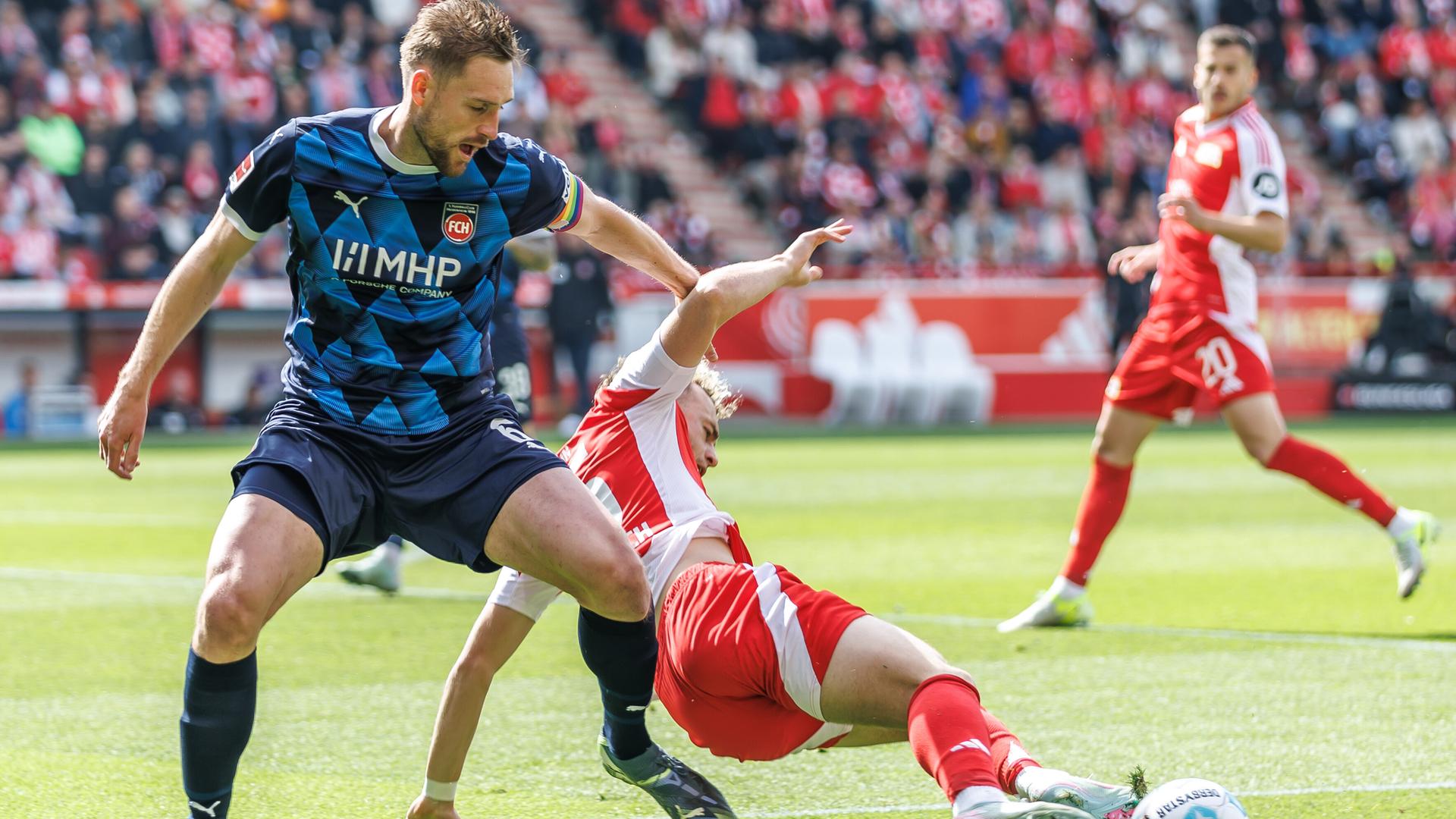 Mainka im blauen Trikot dreht sich auf dem Rasen; Hollerbach im roten Trikot fällt um. Dahinter weitere Spieler und unscharf eine vollbesetzte Zuschauertribüne. Mainka im blauen Trikot dreht sich auf dem Rasen; Hollerbach im roten Trikot fällt um. Dahinter weitere Spieler und unscharf eine vollbesetzte Zuschauertribüne.