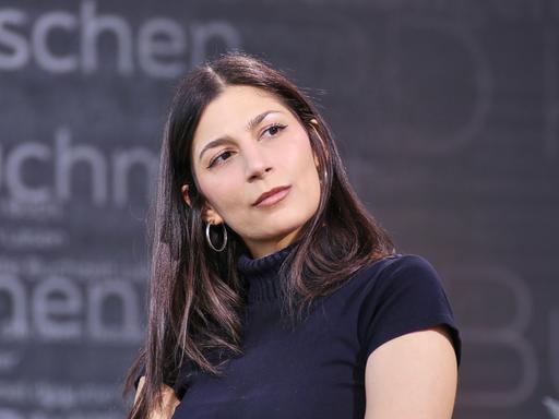 Miriam Davoudvandi trägt ein schwarzes Rollkragen-T-Shirt und hat den Kopf zur Seite geneigt.