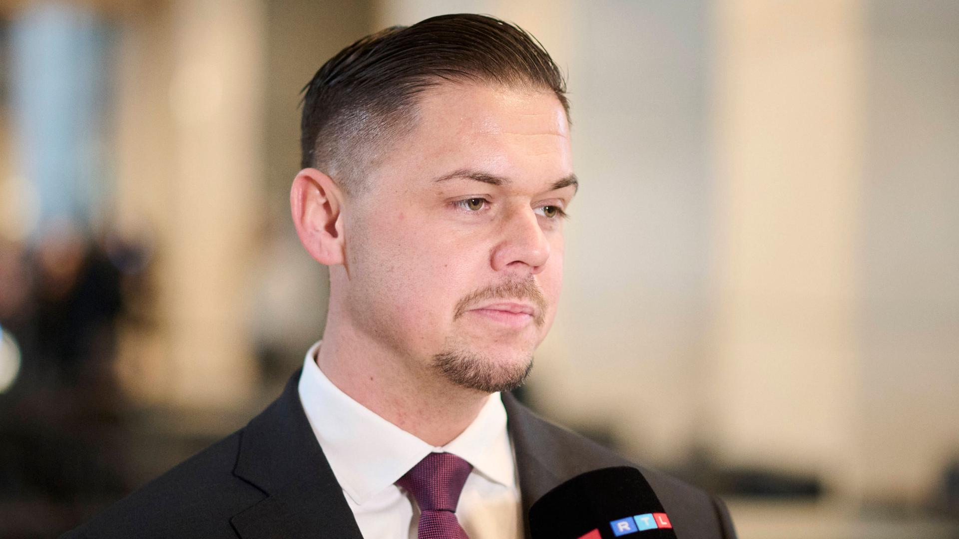 Hannes Gnauck gibt im Bundestag ein Interview. Er hat sich eine Krawatte umgebunden. Sein Undercut ist streng geordnet. Er guckt schläfrig. Außerdem trägt er ein kurz geschnittenes Kinn- und Oberlippenbärtchen. Hannes Gnauck gibt im Bundestag ein Interview. Er hat sich eine Krawatte umgebunden. Sein Undercut ist streng geordnet. Er guckt schläfrig. Außerdem trägt er ein kurz geschnittenes Kinn- und Oberlippenbärtchen.