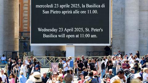 Eine große Videoleinwand verlautbart auf Italienisch und Englisch, dass die Basilika St. Peter am Mittwoch, 23.05.2025 ab 11 Uhr geöffnet sein soll. Im Vordergrund eine Menschenmenge; im Hintergrund die Kolonnaden, die den Petersplatz begrenzen.