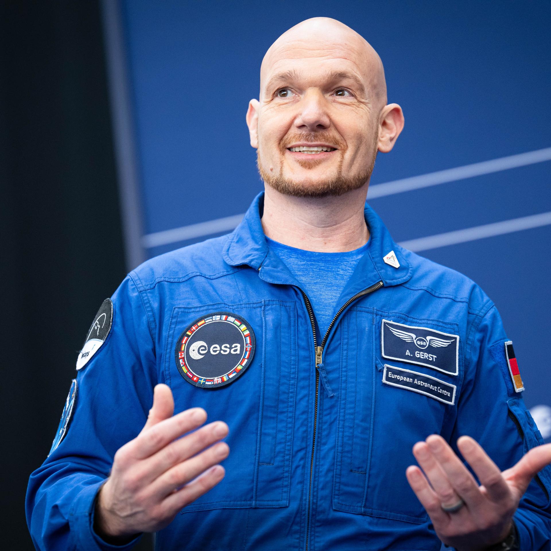Astronaut Alexander Gerst im blauen Overall gestikuliert lächelnd bei der Esa-Ministerratstagung.