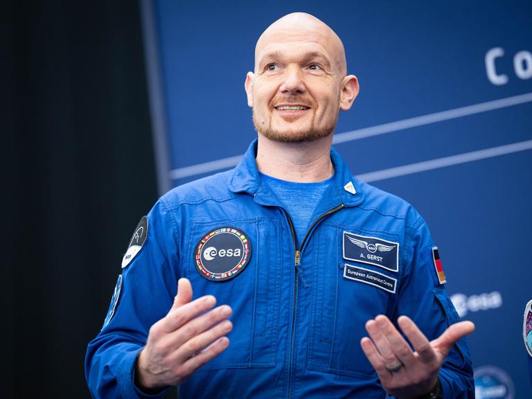Astronaut Alexander Gerst im blauen Overall gestikuliert lächelnd bei der Esa-Ministerratstagung.