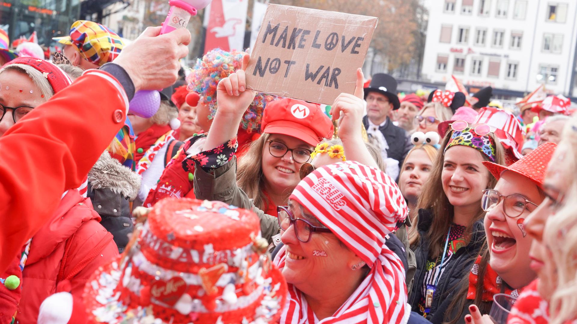 Verkleidete Karnevalisten drängen sich auf dem Kölner Heumarkt. Eine Frau hält ein Schild in die Höhe mit der Aufschrift "Make Love not War".