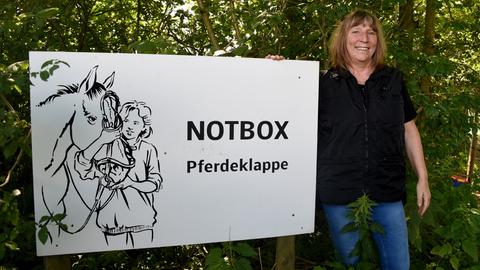 Pferdeklappe-Chefin Petra Teegen steht in Norderbrarup vor einem Schild mit der Aufschrift Notbox.