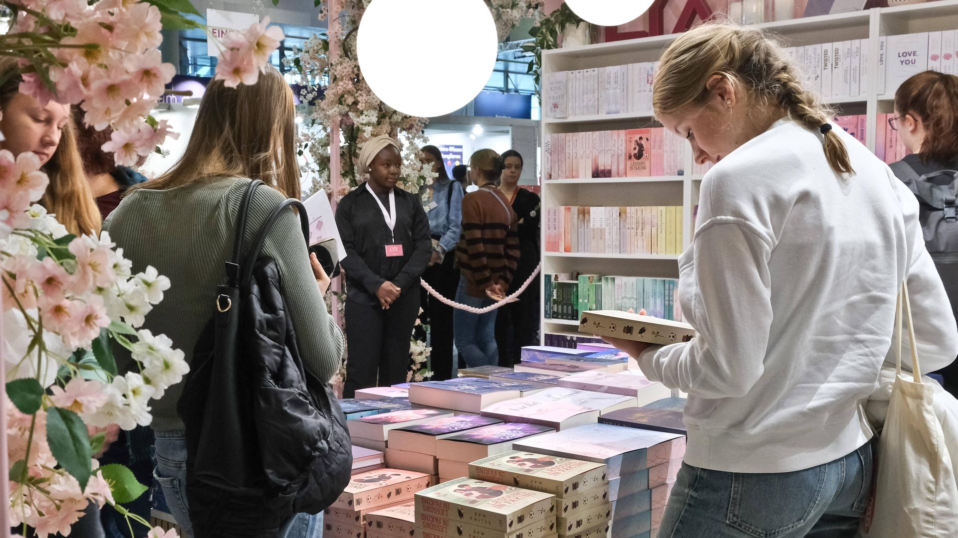 Zwei junge Frauen stehen vor gestapelten Büchern eines Messestands. Die Frau rechts hält ein Buch in der Hand und schaut darauf. Im Hintergrund sieht man weitere Bücherregale und Messebesucher. 