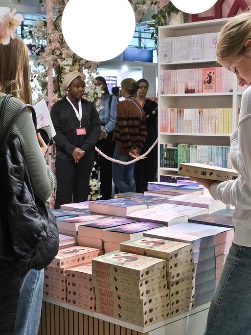 Zwei junge Frauen stehen vor gestapelten Büchern eines Messestands. Die Frau rechts hält ein Buch in der Hand und schaut darauf. Im Hintergrund sieht man weitere Bücherregale und Messebesucher. 