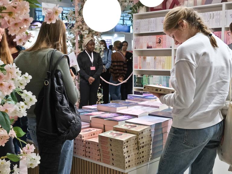 Zwei junge Frauen stehen vor gestapelten Büchern eines Messestands. Die Frau rechts hält ein Buch in der Hand und schaut darauf. Im Hintergrund sieht man weitere Bücherregale und Messebesucher. 