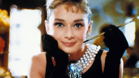 Audrey Hepburn sitzt in edler Kleidung an einem Tisch in einem Luxus-Restaurant und lächelt freundlich in die Kamera.