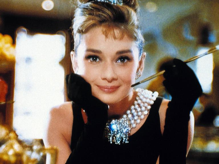 Audrey Hepburn sitzt in edler Kleidung an einem Tisch in einem Luxus-Restaurant und lächelt freundlich in die Kamera.