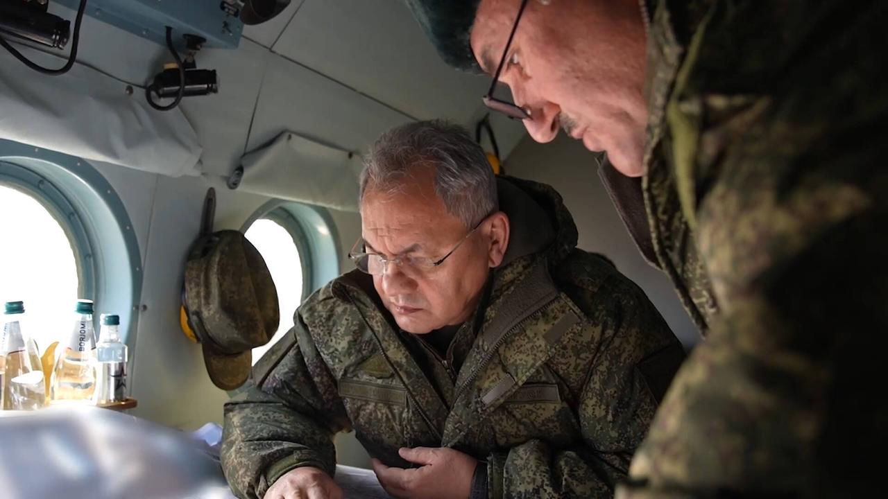 Russlands Verteidigungsminister trägt Tarnkleidung und schaut in einem Flugzeug auf etwas herunter. Neben ihm steht ein Soldat.