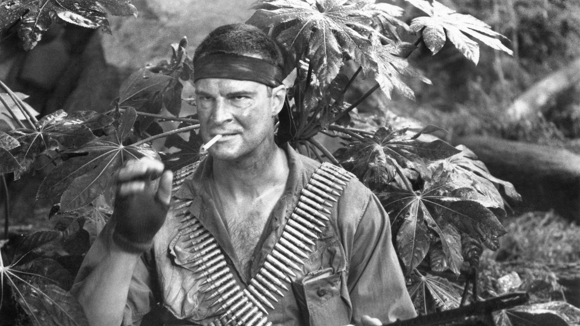 Der Schauspieler Richard Moll zündet sich im Dschungel eine Zigarette an. Er trägt einen Patronengurt und ein Maschinengewehr.