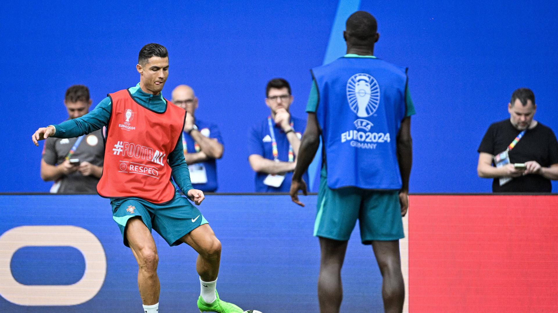 Cristiano Ronaldo steht auf einem Fußballplatz und passt einen Ball zu einem Mitspieler, der mit dem Rücken zur Kamera steht. Ronaldo trägt ein rotes Trainingsleibchen über seinem blauen Trikot. Er ist kurz davor, den ball mit seinem Fuß zu berühren. Er streckt konzentriert die Zunge heraus. 