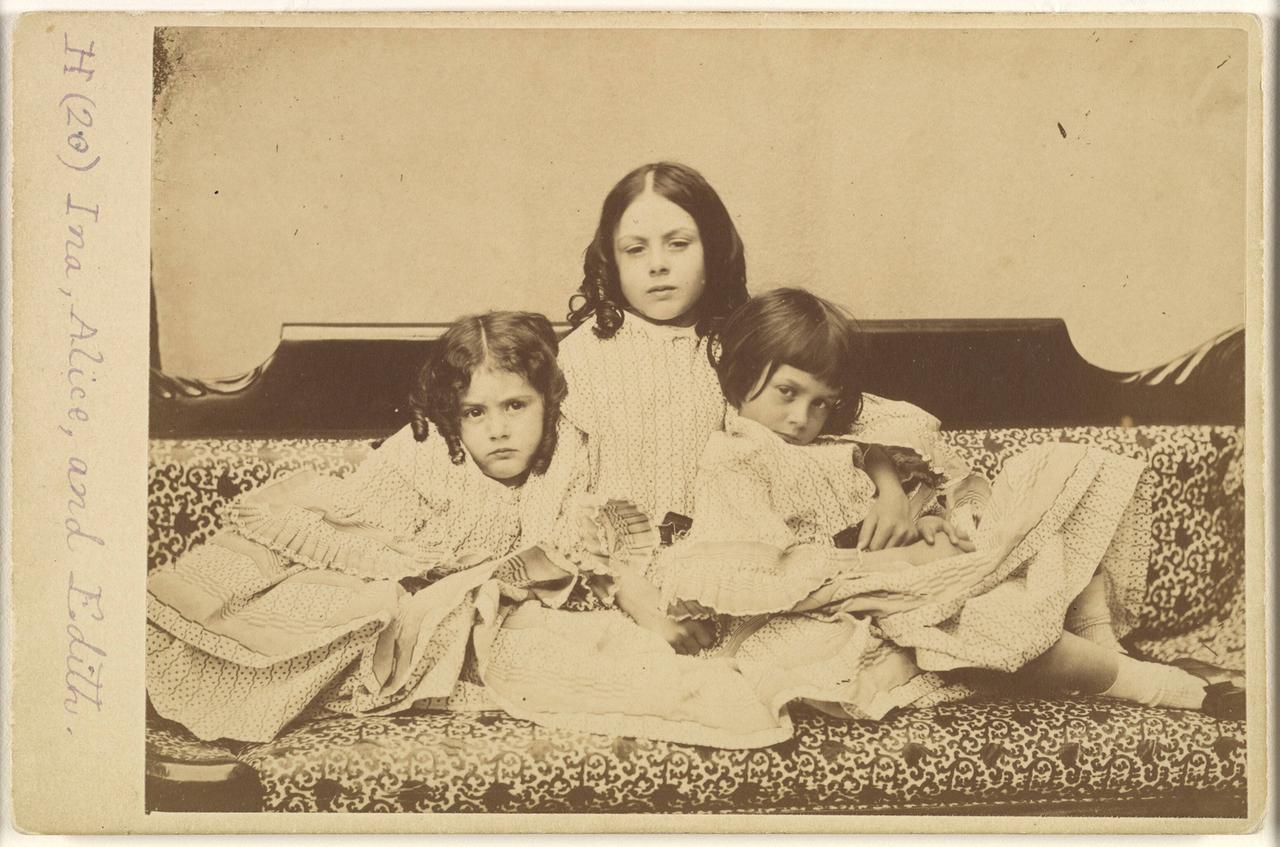Historische Aufnahme (in Sepia-Tönung) der Geschwister Edith, Ina und Alice Liddell auf einem Sofa im Sommer 1858.