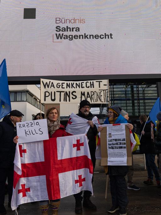 Demonstranten stehen mit einem Schild "Wagenknecht - Putins Marionette" und einer georgischen Fahne vor dem World Conference Center Bonn, der mit dem Logo des BSW geschmückt ist.