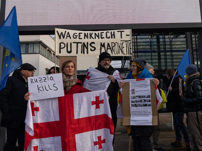 Demonstranten stehen mit einem Schild "Wagenknecht - Putins Marionette" und einer georgischen Fahne vor dem World Conference Center Bonn, der mit dem Logo des BSW geschmückt ist.