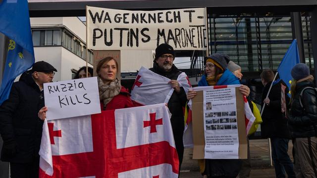 Demonstranten stehen mit einem Schild "Wagenknecht - Putins Marionette" und einer georgischen Fahne vor dem World Conference Center Bonn, der mit dem Logo des BSW geschmückt ist.