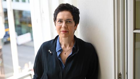 Sonja Anders trägt eine dunkle Jacke und lehnt an einer weißen Wand in den Räumlichkeiten des Theaters. Sie schaut freundlich und direkt in die Kamera.