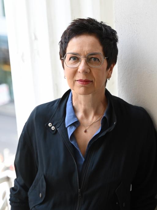 Sonja Anders trägt eine dunkle Jacke und lehnt an einer weißen Wand in den Räumlichkeiten des Theaters. Sie schaut freundlich und direkt in die Kamera.