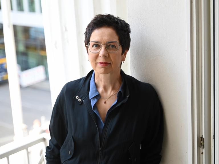 Sonja Anders trägt eine dunkle Jacke und lehnt an einer weißen Wand in den Räumlichkeiten des Theaters. Sie schaut freundlich und direkt in die Kamera.