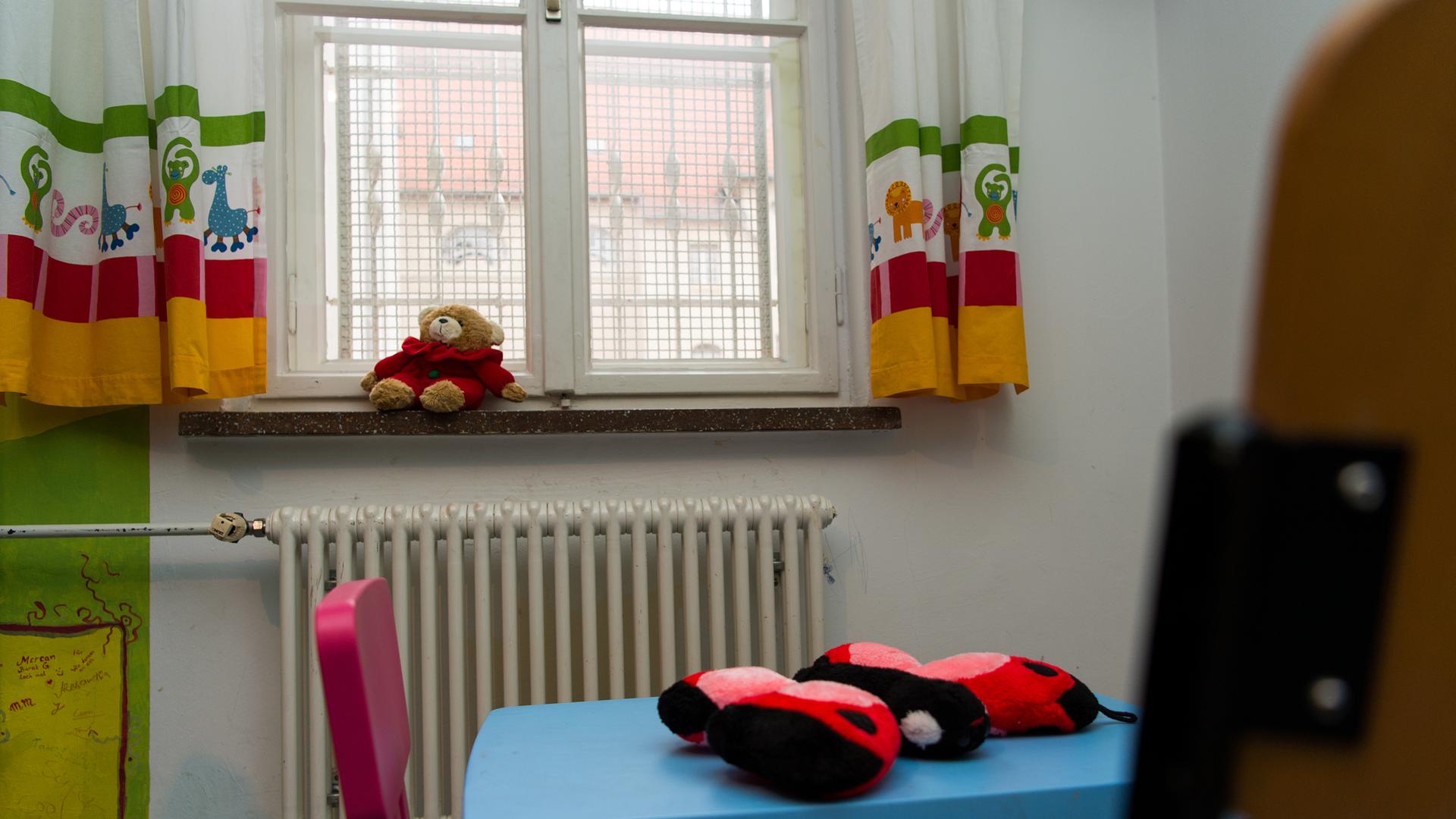 Vor einem vergitterten Fenster liegt auf der Fensterbank ein Teddybär. Im Raum stehen bunte Kindermöbel.