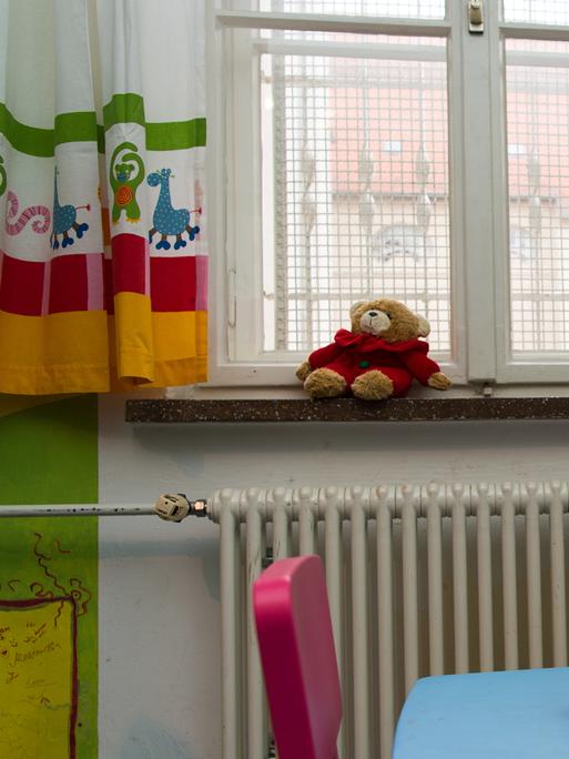 Vor einem vergitterten Fenster liegt auf der Fensterbank ein Teddybär. Im Raum stehen bunte Kindermöbel.