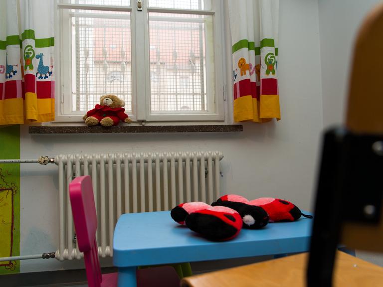 Vor einem vergitterten Fenster liegt auf der Fensterbank ein Teddybär. Im Raum stehen bunte Kindermöbel.