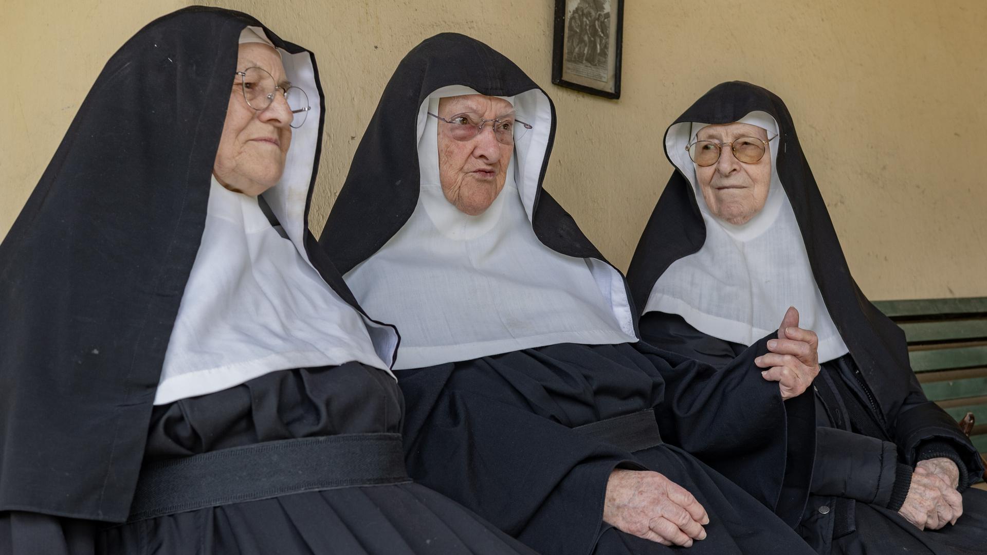 Drei alte Frauen in Ordensornat (Schwester Rita, Schwester Bernadette, Schwester Regina sitzen nebeneinander auf einer Bank Drei alte Frauen in Ordensornat (Schwester Rita, Schwester Bernadette, Schwester Regina sitzen nebeneinander auf einer Bank
