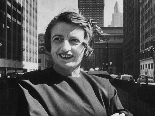 Porträt in Schwarzweiß von Ayn Rand, lächelnd und mit verschränkten Armen vor dem Grand Central Gebäude in Midtown Manhattan.