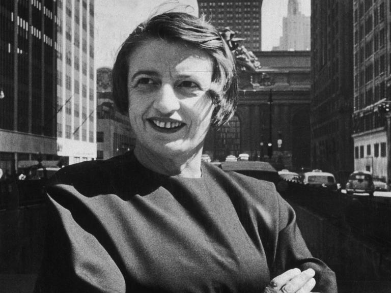 Porträt in Schwarzweiß von Ayn Rand, lächelnd und mit verschränkten Armen vor dem Grand Central Gebäude in Midtown Manhattan.