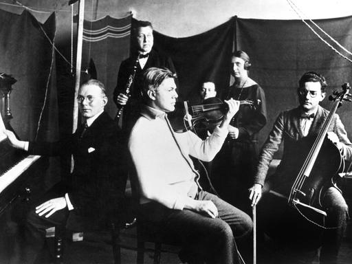 Schwarz-Weiß-Fotografie eines Kammermusikensembles, das für einen Radiosender in den 1920er-Jahren musiziert. Im Hintergrund sind eine Frau mit Kopfhörern und ein Mann zu sehen.
