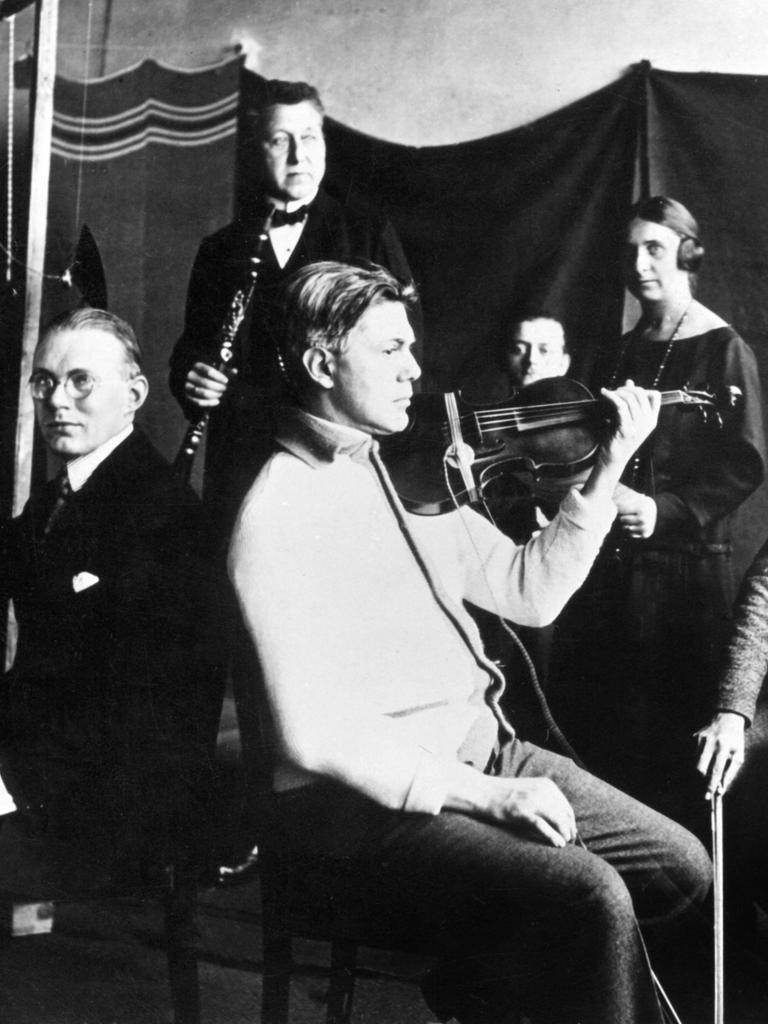 Schwarz-Weiß-Fotografie eines Kammermusikensembles, das für einen Radiosender in den 1920er-Jahren musiziert. Im Hintergrund sind eine Frau mit Kopfhörern und ein Mann zu sehen.