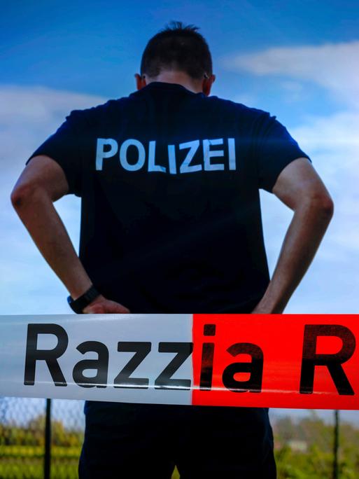 Ein Polizist steht vor blauem Himmel mit dem Rücken zur Kamera. Davor Absperrband mit der Aufschrift Razzia.