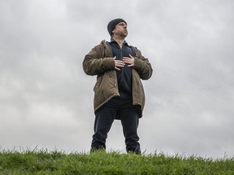 Bjarne Mädel als Kommissar Sörensen steht auf einem Deich, blickt nach oben und hält die Hände auf die Brust.