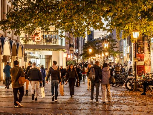 Passanten laufen durch die Fußgängerzone von München bei beginnender Abenddämmerung. Man sieht Straßenlaternen und Geschäfte, im Vordergrund ein Baum.