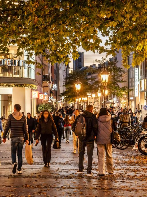 Passanten laufen durch die Fußgängerzone von München bei beginnender Abenddämmerung. Man sieht Straßenlaternen und Geschäfte, im Vordergrund ein Baum. Passanten laufen durch die Fußgängerzone von München bei beginnender Abenddämmerung. Man sieht Straßenlaternen und Geschäfte, im Vordergrund ein Baum.