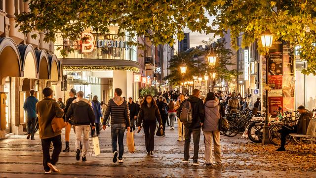 Passanten laufen durch die Fußgängerzone von München bei beginnender Abenddämmerung. Man sieht Straßenlaternen und Geschäfte, im Vordergrund ein Baum.