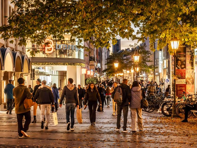 Passanten laufen durch die Fußgängerzone von München bei beginnender Abenddämmerung. Man sieht Straßenlaternen und Geschäfte, im Vordergrund ein Baum. Passanten laufen durch die Fußgängerzone von München bei beginnender Abenddämmerung. Man sieht Straßenlaternen und Geschäfte, im Vordergrund ein Baum.