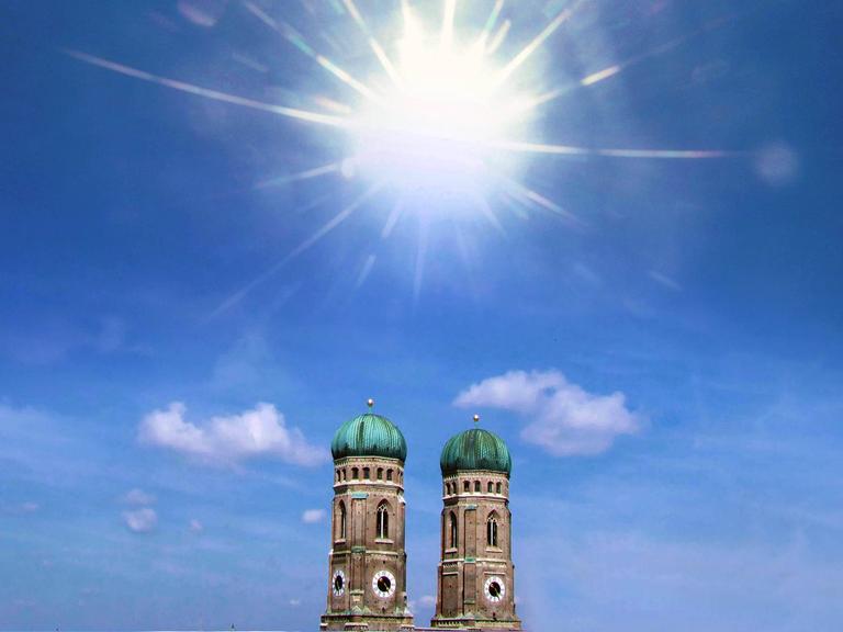 Die Türme der Frauenkirche in München vor blauem Himmel und viel Sonnenschein. Es wirkt sehr heiß.