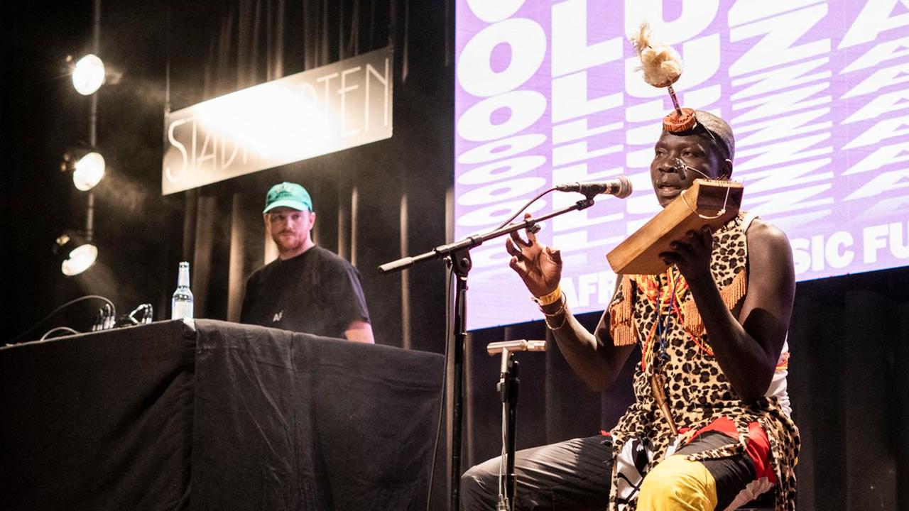 Ein afrikanischer Mann in traditioneller Kleidung sitzt auf der Bühne neben einem DJ und spielt ein in Afrika gebautes Instrument aus Holz.