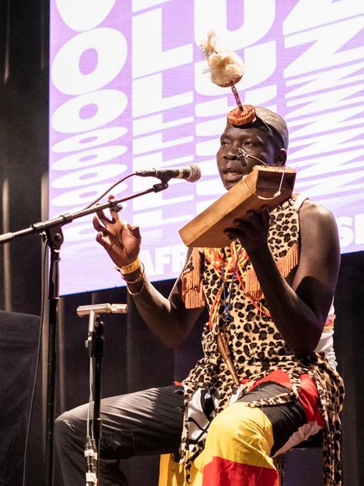 Ein afrikanischer Mann in traditioneller Kleidung sitzt auf der Bühne neben einem DJ und spielt ein in Afrika gebautes Instrument aus Holz.