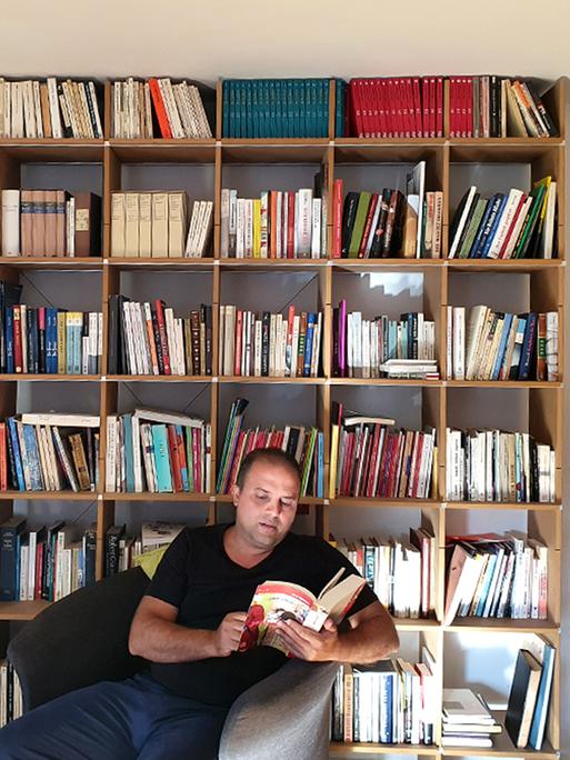 Der jemenitische Dichter Osama Al-Dhari sitzt vor einem Bücherregel und blätter in einem Buch. 