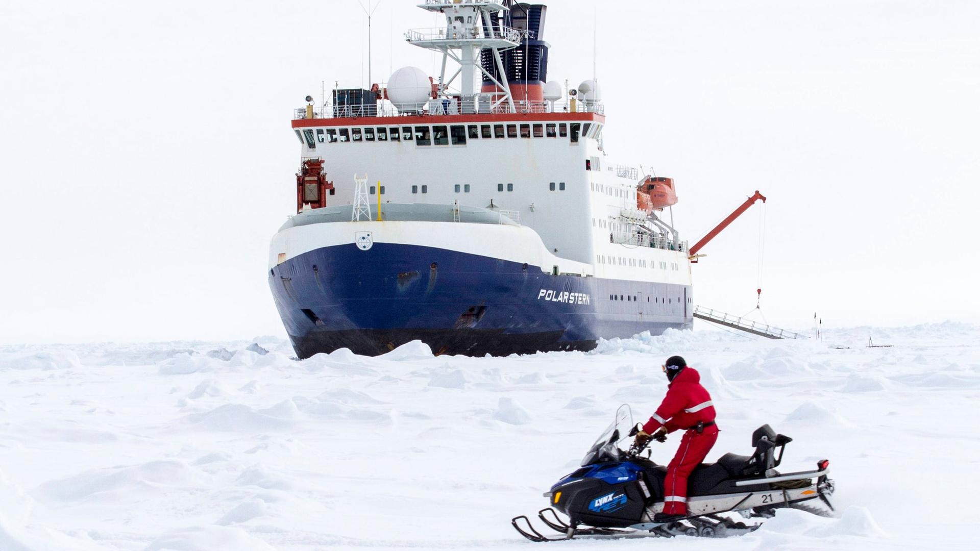 Das Bild zeigt den Forschungseisbrecher Polarstern, der von Eisschollen umgeben ist. Im Vordergrund ist ein Forscher auf einem Schneemobil zu sehen.  