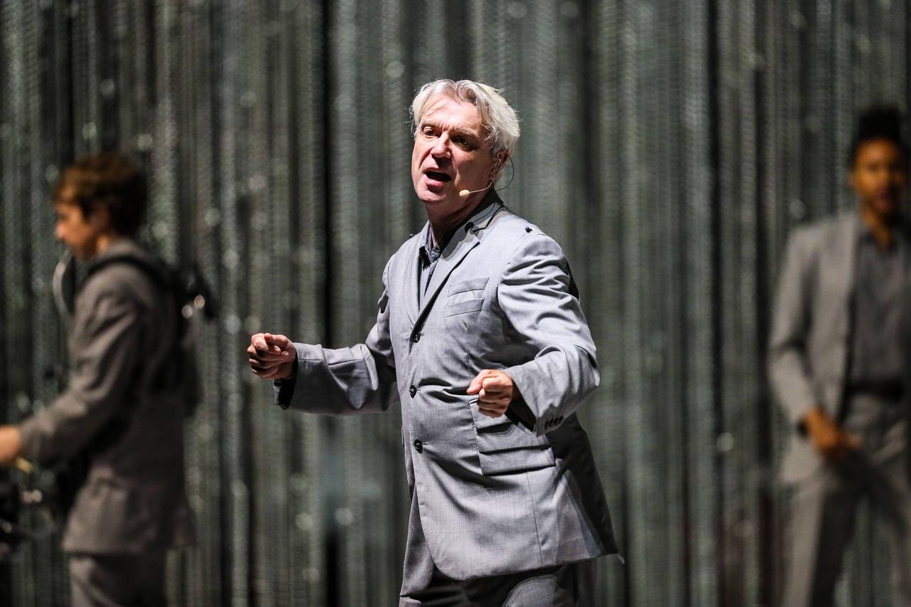 David Byrne steht auf der Bühne und bewegt sich. Er hat weiße Haare, trägt einen silbergrauen Anzug. Im Hintergrund ein dunkelgrauer Vorhang.