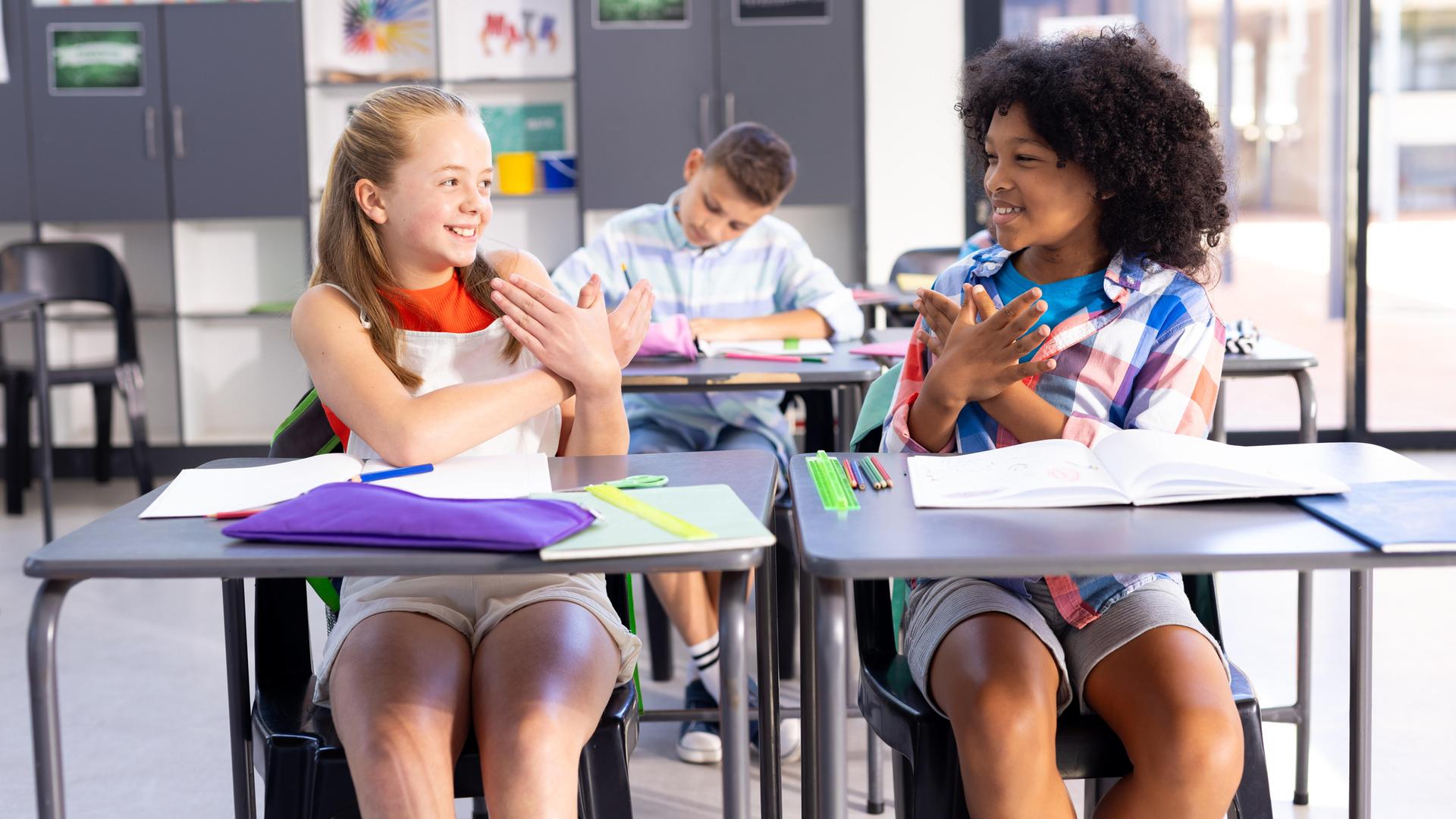 Zwei lächelnde Mädchen unterhalten sich im Klassenraum in Gebärdensprache