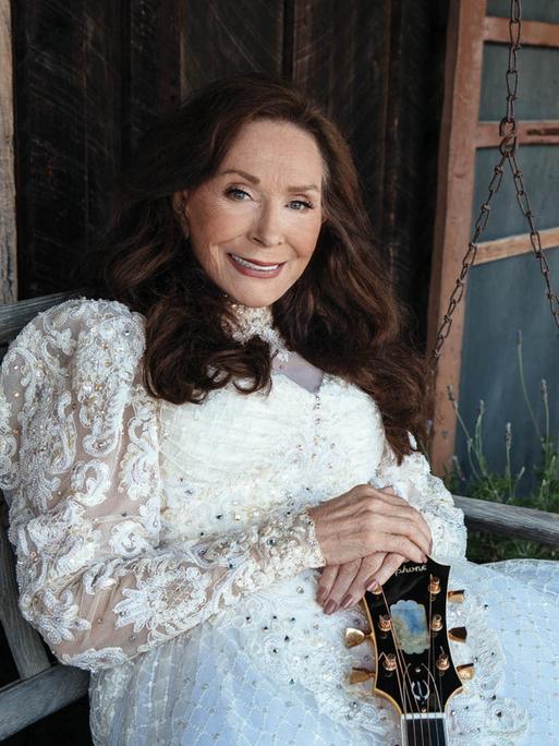 Countrylegende Loretta Lynn in einem weißen Kleid vor einem Holzhaus.