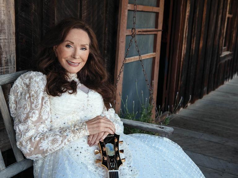 Countrylegende Loretta Lynn in einem weißen Kleid vor einem Holzhaus.