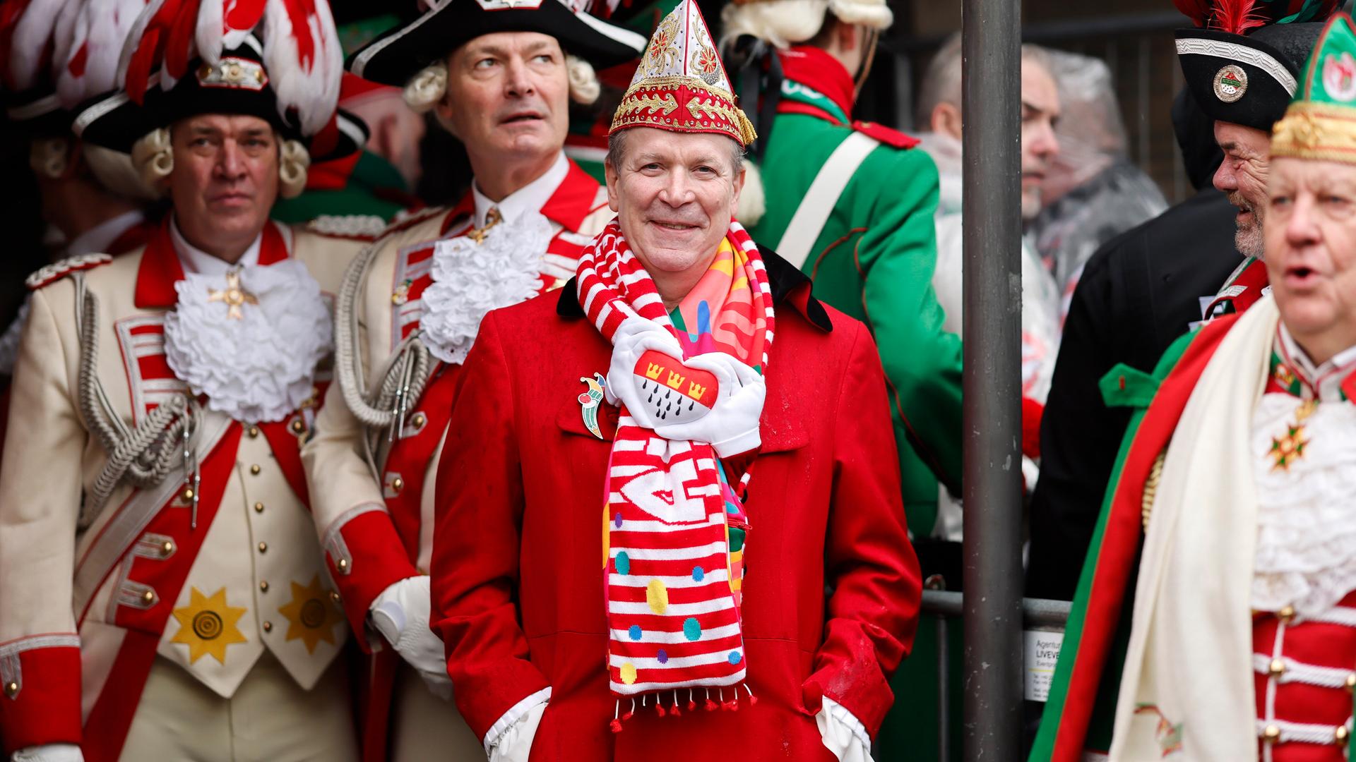 Karnevalist Christoph Kuckelkorn schaut in Uniform in die Kamera und lächelt, hinter ihm stehen weitere Personen in Festuniform. Karnevalist Christoph Kuckelkorn schaut in Uniform in die Kamera und lächelt, hinter ihm stehen weitere Personen in Festuniform.